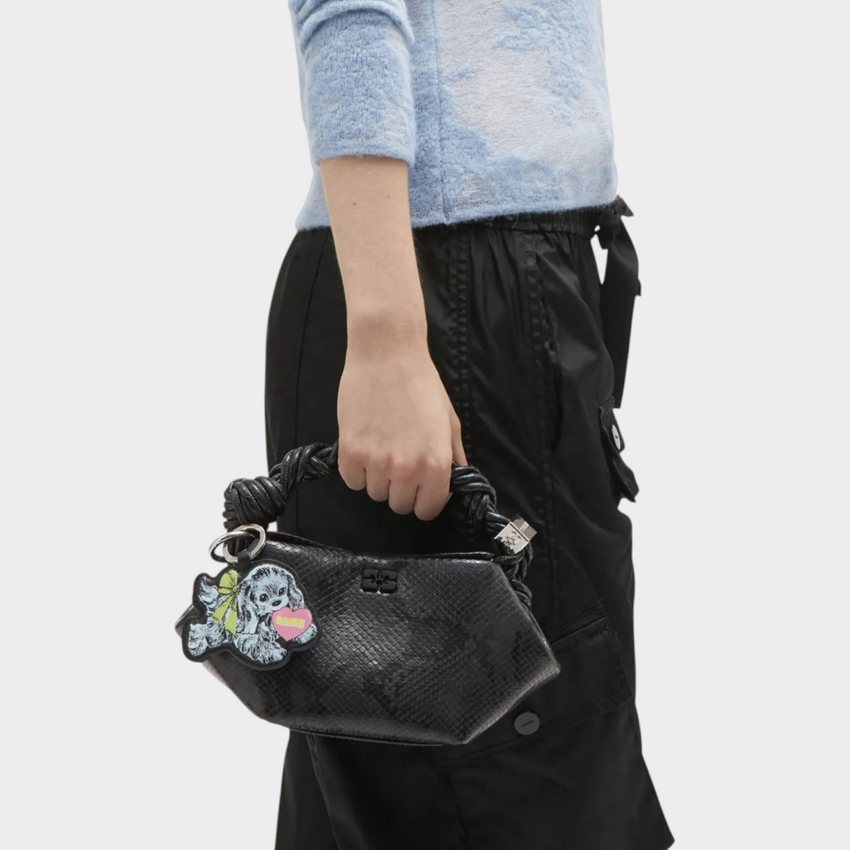 Supplied Fashion Ganni baby blue puppy keyring with dog motif attached to a black snakeskin handbag held by a model