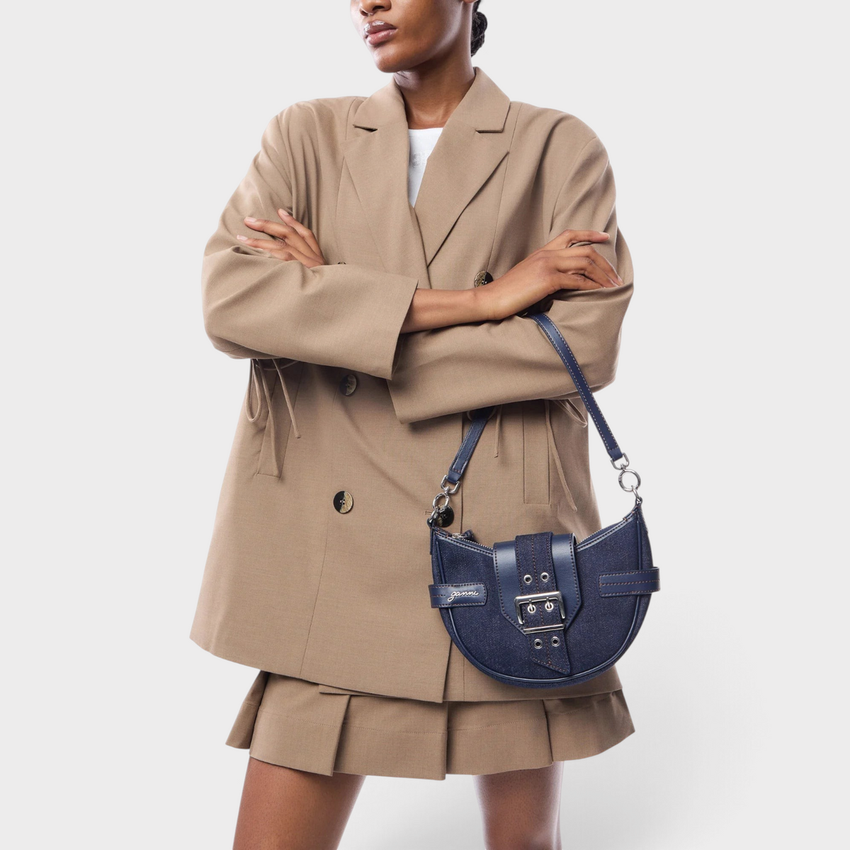 Supplied Fashion GANNI Bucky small blue denim crossbody bag with silver buckle detail worn by a model in a beige suit