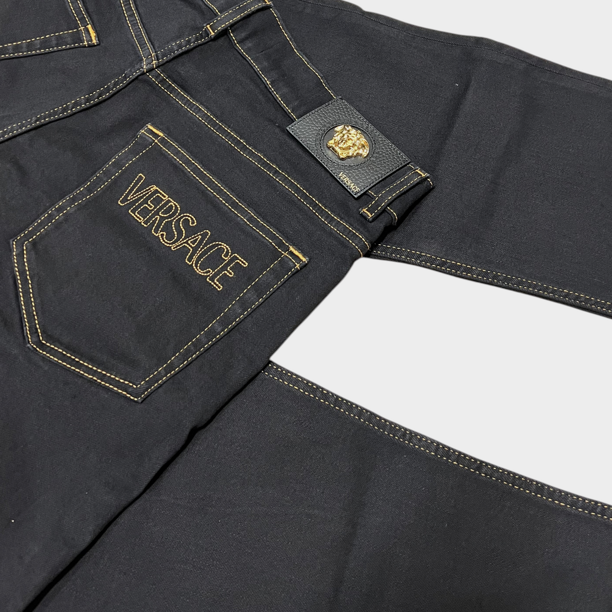 Supplied Fashion black stretch denim jeans with gold stitching, Versace logo embroidery, and Medusa head leather patch.