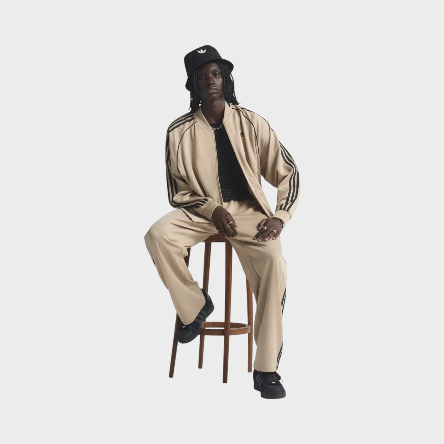 Supplied Fashion model wearing a beige and black loose track top with iconic three stripes, seated on a wooden stool.
