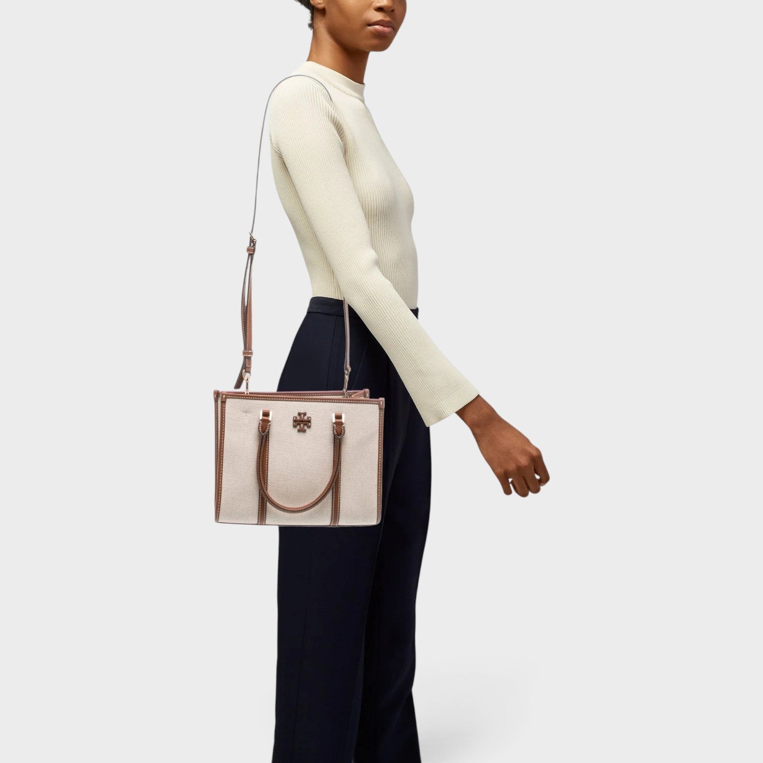 Supplied Fashion beige canvas top-handle bag with brown leather trim carried by a model in a cream top and trousers.