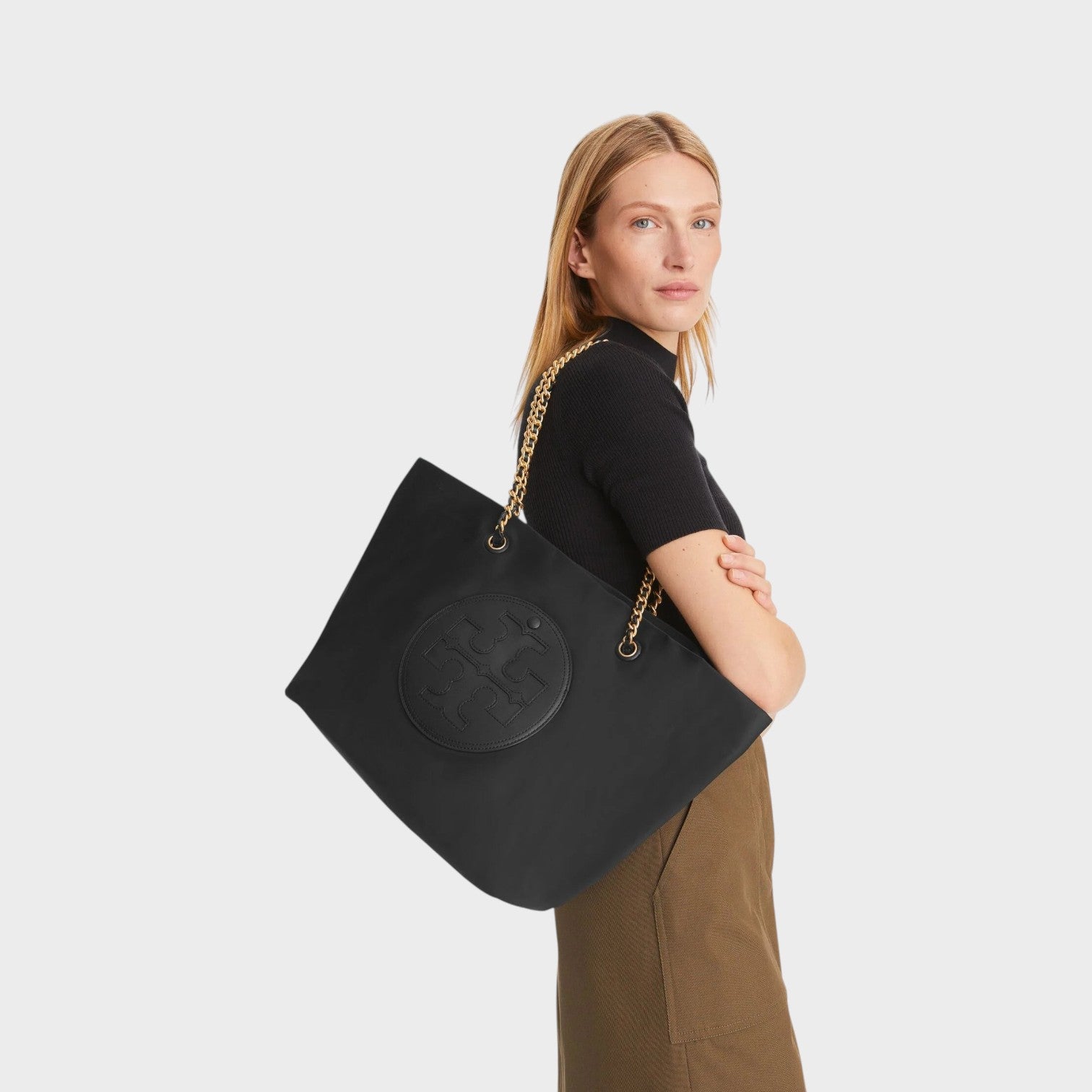 Model carrying a black Tory Burch Ella chain tote bag with gold hardware on her shoulder, Supplied Fashion.