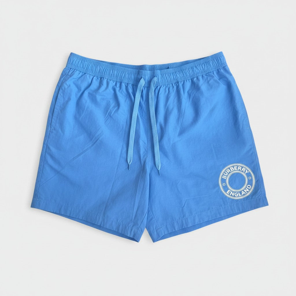 Supplied Fashion periwinkle blue nylon swim shorts with a circular white logo on the leg and an elastic drawstring.